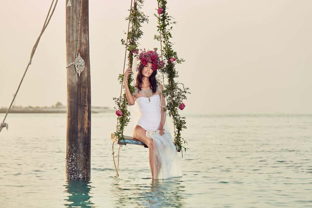 Bride sitting on a floral swing over water, wearing a white wedding dress and flower crown, with a s.