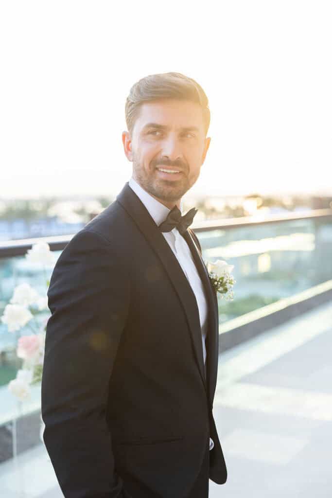 Groom in classic black tuxedo with bow tie, smiling outdoors at waterfront wedding venue.