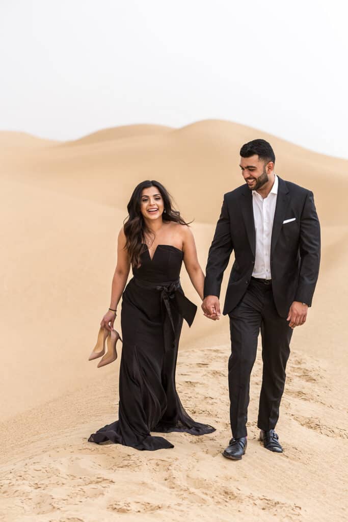 A couple in Blakc tie outfit walks the desert in dubai during their engagement session.
