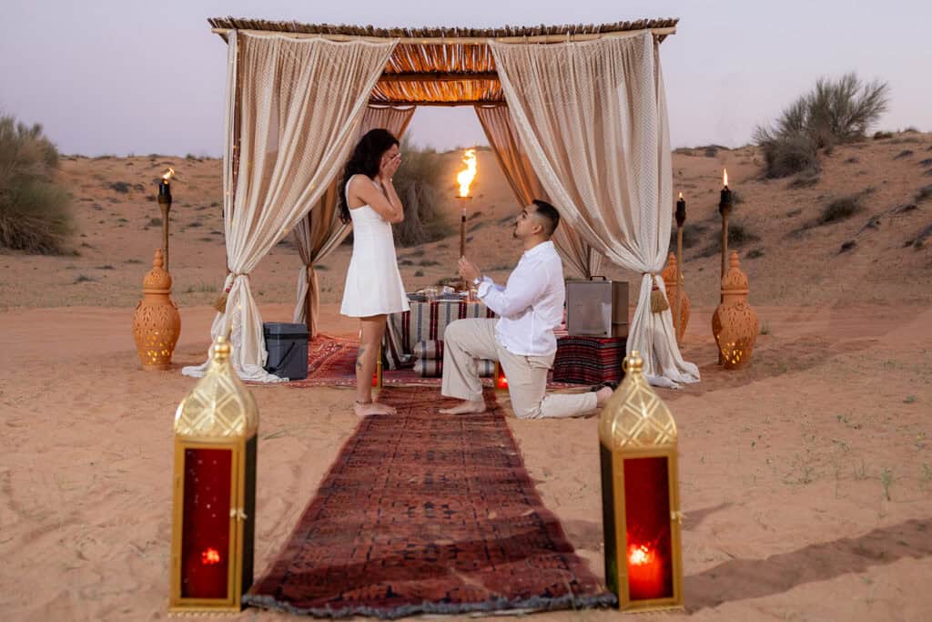 A young man kneels down and propose to his girlfriend in an outdoor desert setting at sunset.